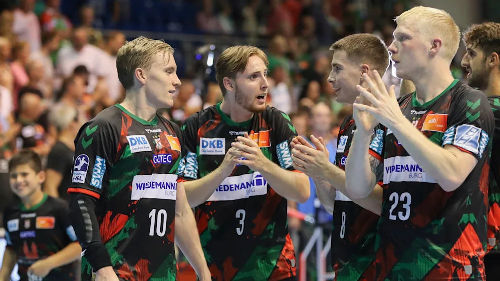 Handball players in black and red uniforms celebrating on a court with spectators in the background.