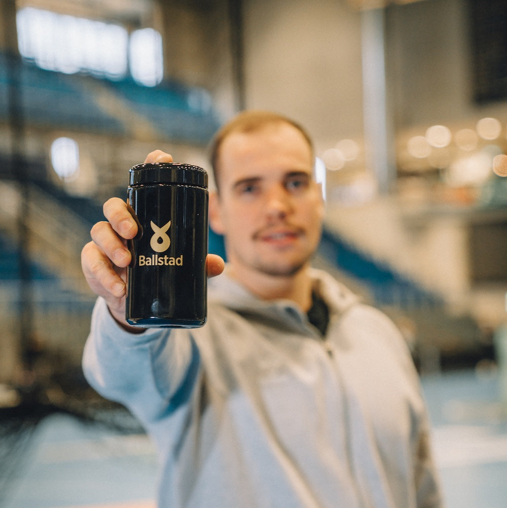 Person holding a black container with 'Ballstad' branding in an indoor setting.