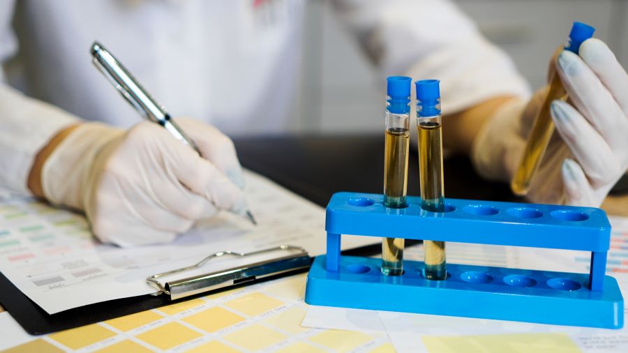 A laboratory technician wearing white gloves records data on a clipboard while handling test tubes filled with yellow liquid, placed in a blue test tube rack.