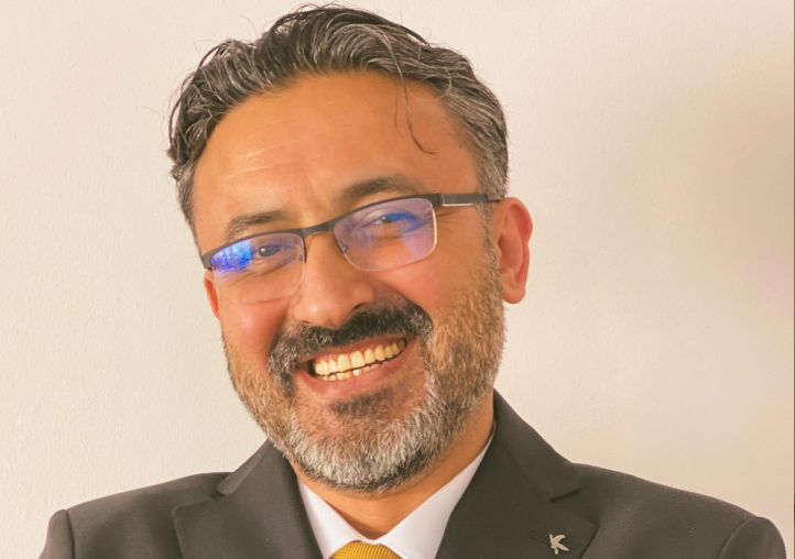Smiling man with short dark hair and gray beard, wearing glasses, a black suit jacket, white shirt, and yellow tie, standing against a plain light background.