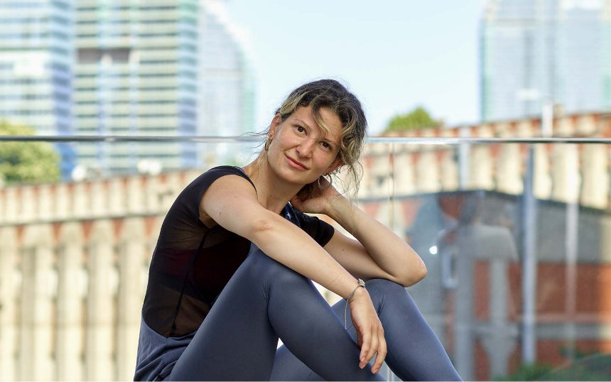 A smiling young woman with curly hair pulled back is sitting outdoors on a rooftop or elevated urban setting, leaning slightly with one hand resting on the back of her head. 