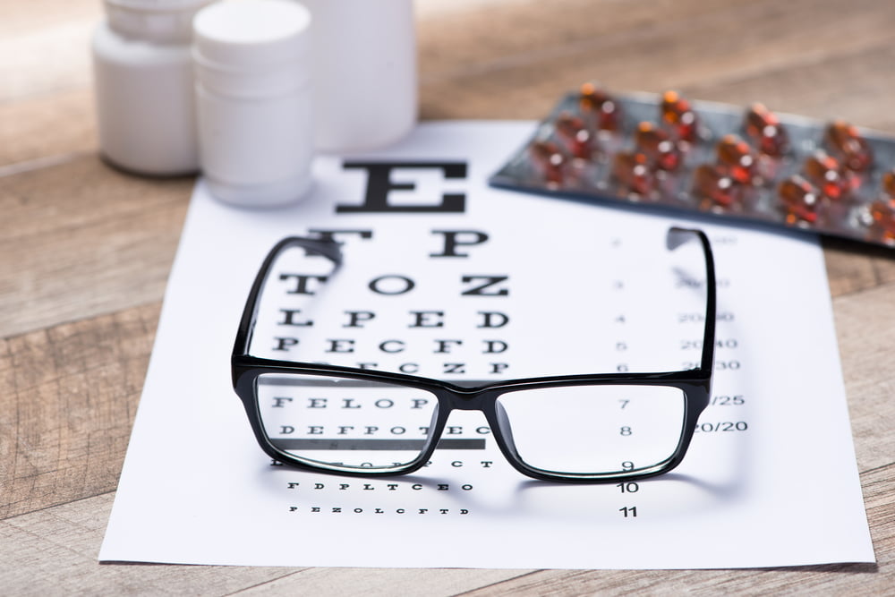 Eyeglasses resting on a vision test chart with medicine bottles and a blister pack of capsules in the background, representing eye health and vision care.