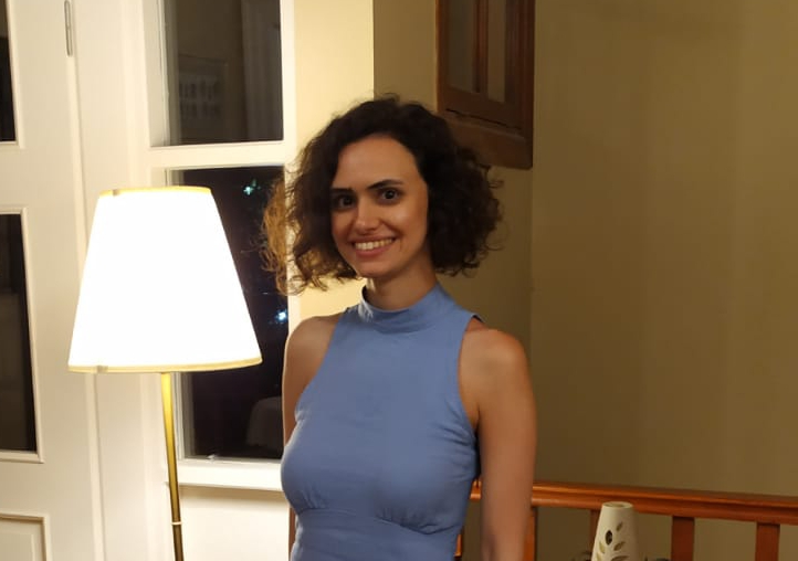 Smiling woman with curly dark hair wearing a sleeveless blue dress, standing indoors near a lit floor lamp and a window at night.