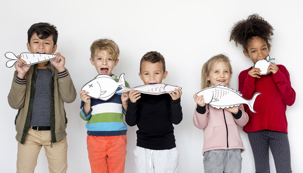 A group of five diverse children standing in a row, smiling and playfully holding illustrated paper fish cutouts in front of their mouths, representing the importance of fish in a healthy diet.