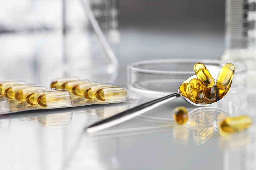 Close-up of omega-3 fish oil capsules on a metal spoon, with more capsules in blister packs and laboratory glassware in the background, symbolizing scientific formulation and supplement quality.