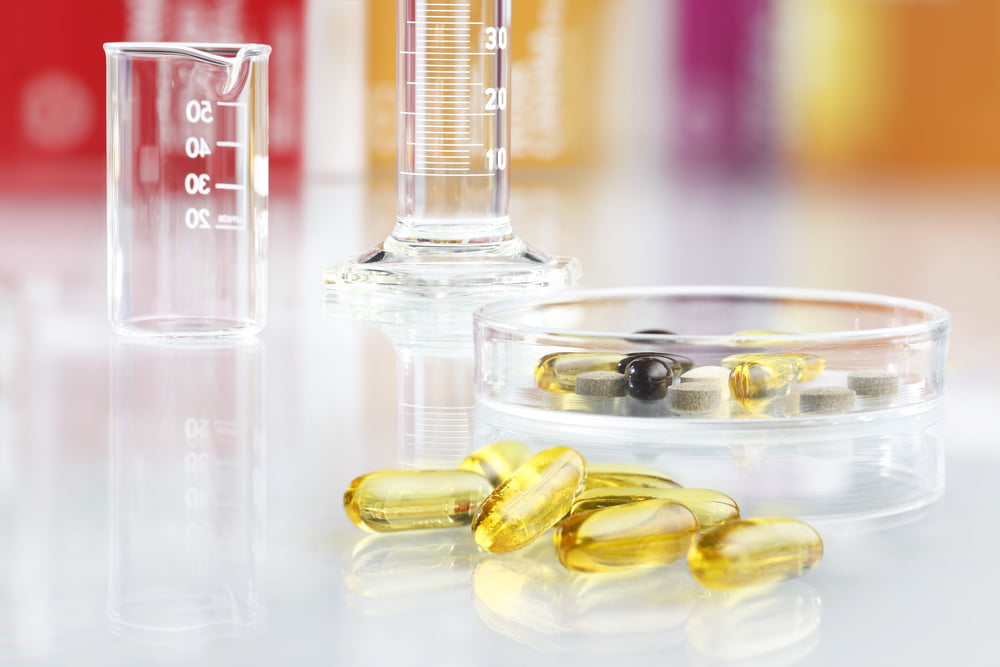 Close-up of omega-3 fish oil capsules on a laboratory surface with a petri dish, beaker, and graduated cylinder in the background, representing supplement testing and scientific research.