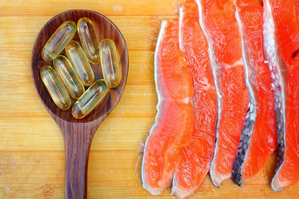 Close-up of fresh salmon fillets arranged on a wooden cutting board next to a wooden spoon filled with golden omega-3 fish oil capsules.