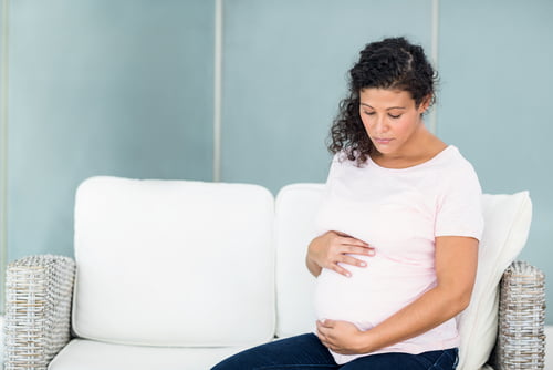 Pregnant woman sitting on a white sofa, gently holding her belly and looking down with a thoughtful expression.