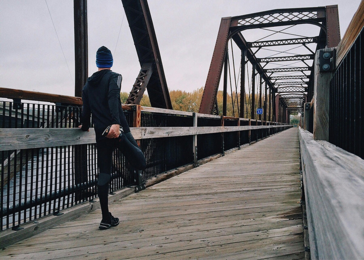 A person wearing black athletic clothes and a blue striped beanie is performing a quad stretch while standing on a wide wooden pedestrian bridge.