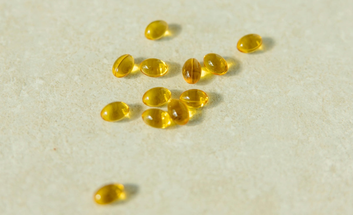 A small pile of shiny, translucent gold soft gel capsules (likely fish oil or omega-3 supplements) is centered in the foreground against a white background.