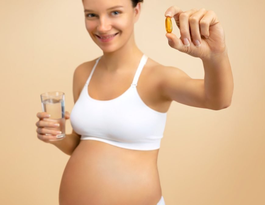 A smiling, pregnant woman wearing a white sports bra and underwear stands against a plain beige background. She holds a small, translucent gold soft gel capsule.