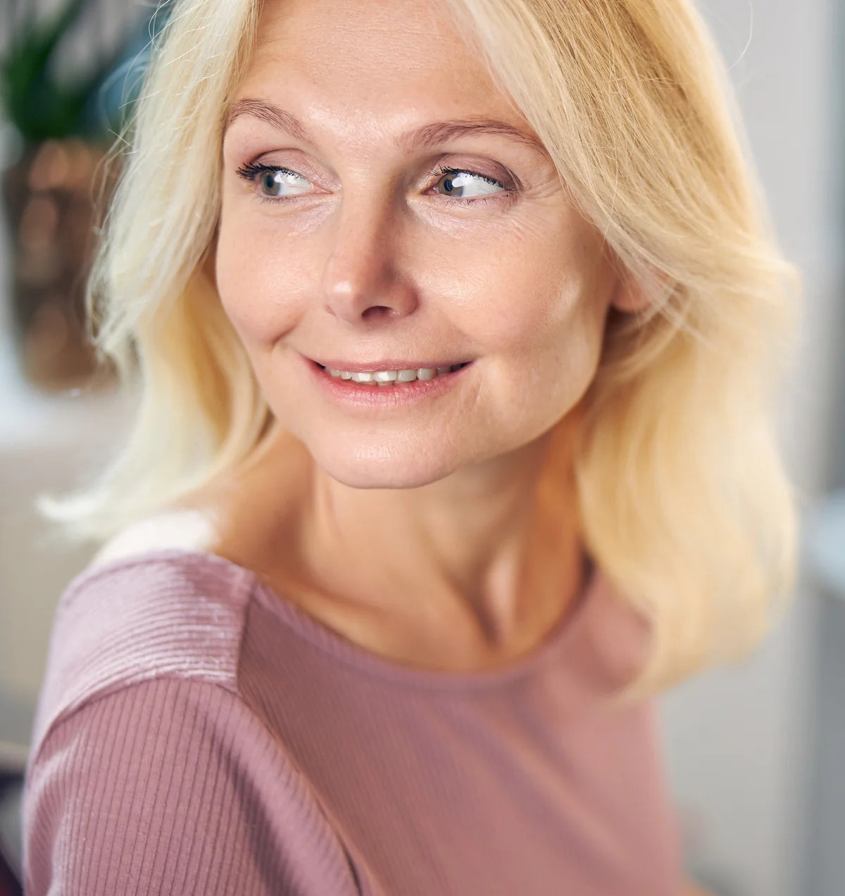 Portrait of a middle-aged blonde woman in a pink top smiling softly while looking to the side.